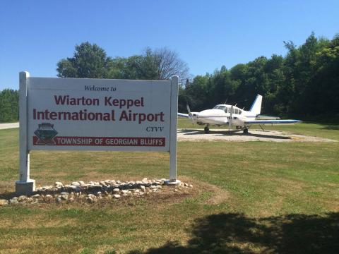 Sign reading Wiarton Keppel International Airport. There is a small airplane behind the sign with two props.