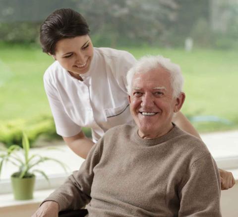 A long term care resident sits in a wheel chair next to a window and is assisted by a healthcare professional