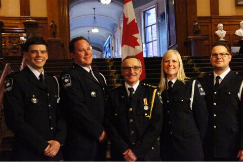 Five Grey County paramedics wearing their formal uniforms stand in front of a Canadian flag inside queen's park in Toronto.